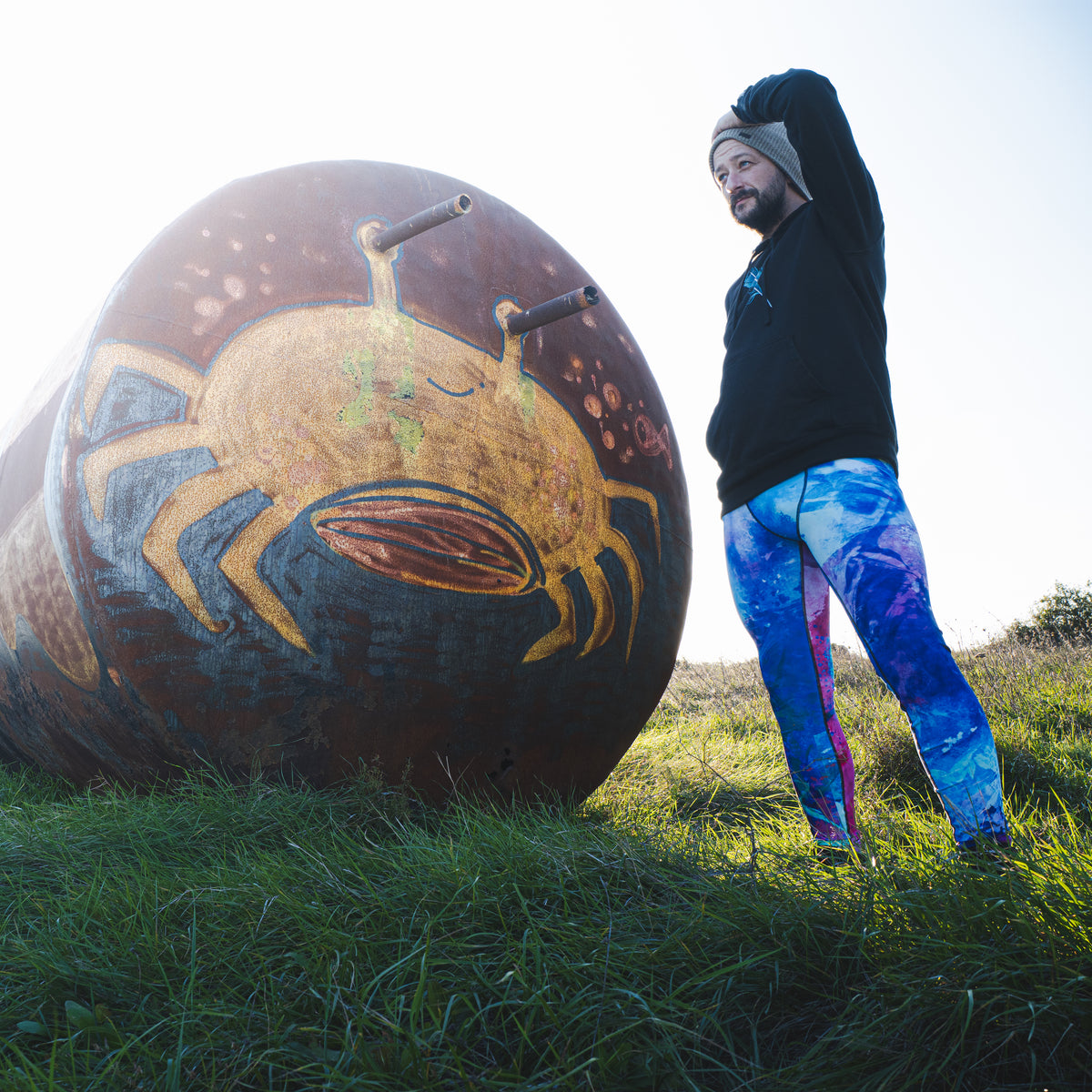 Herren-Leggings Energy Flow von Bachblatt mit blau-grün-violettem Muster – Am Model neben einer bemalten Kugel im Freien, Rückansicht.
