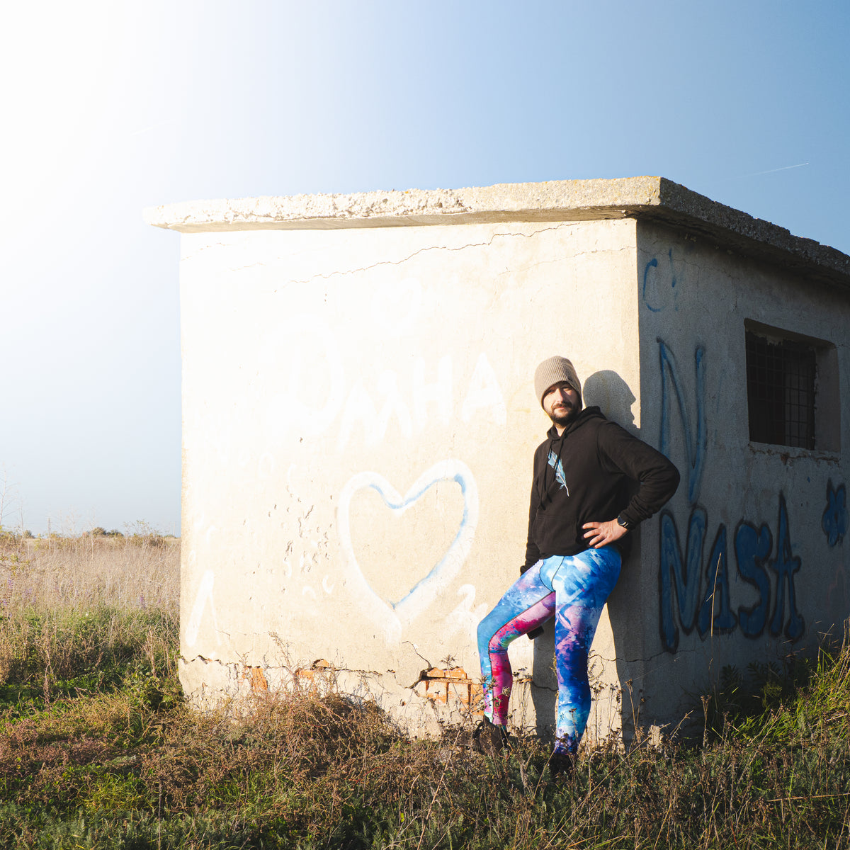 Herren-Leggings Energy Flow von Bachblatt mit blau-grün-violettem Muster – Am Model vor einem kleinen Gebäude mit Graffiti.
